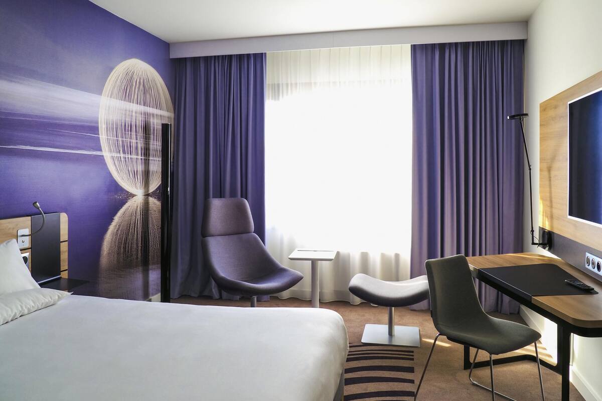 A hotel room with a bed, two chairs, a desk, a TV, and a large window with curtains.