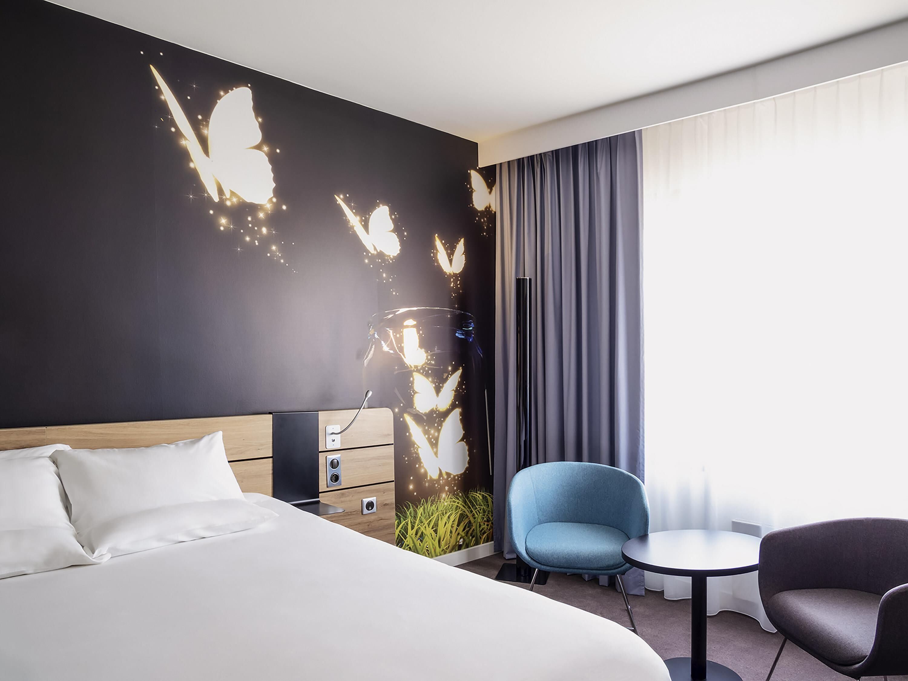 Une chambre d’hôtel avec un lit, un bureau, une chaise bleue, une chaise grise et une grande fenêtre avec des rideaux.