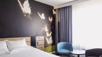 Une chambre d’hôtel avec un lit, un bureau, une chaise bleue, une chaise grise et une grande fenêtre avec des rideaux.