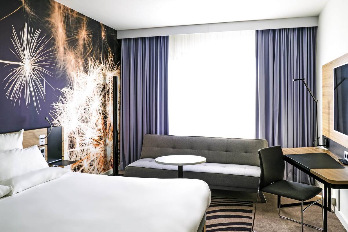 A hotel room with a bed, a sofa, a desk, and a large window with curtains.