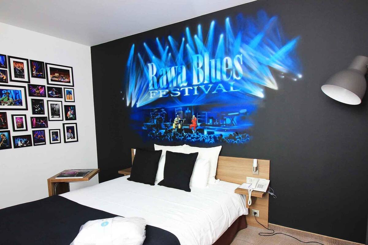 A hotel room with a bed, a nightstand, a phone, and a wall with framed photos.