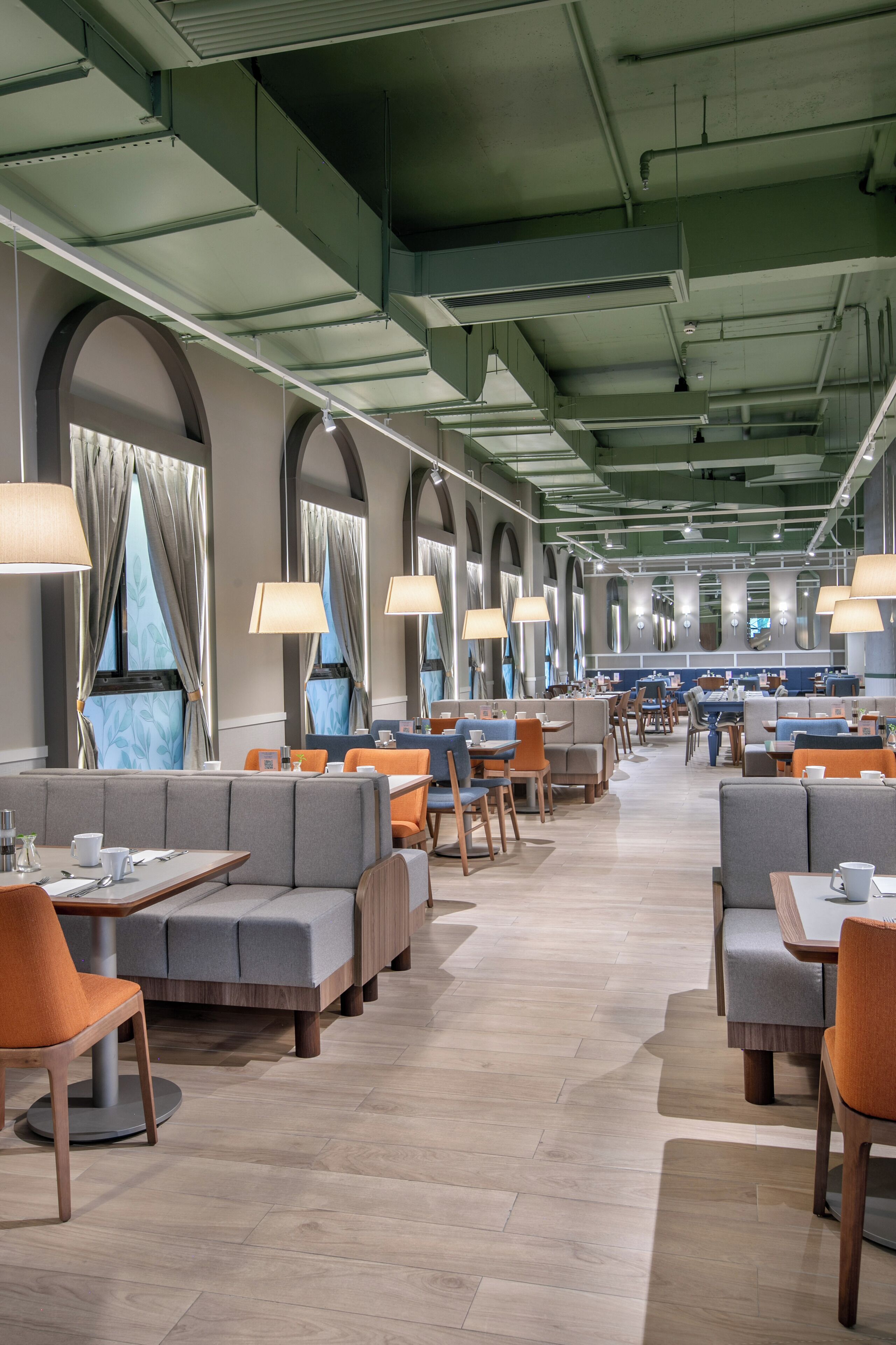 Um restaurante espaçoso com pé-direito alto, grandes janelas com cortinas e diversas opções de assentos, incluindo cabines e mesas.