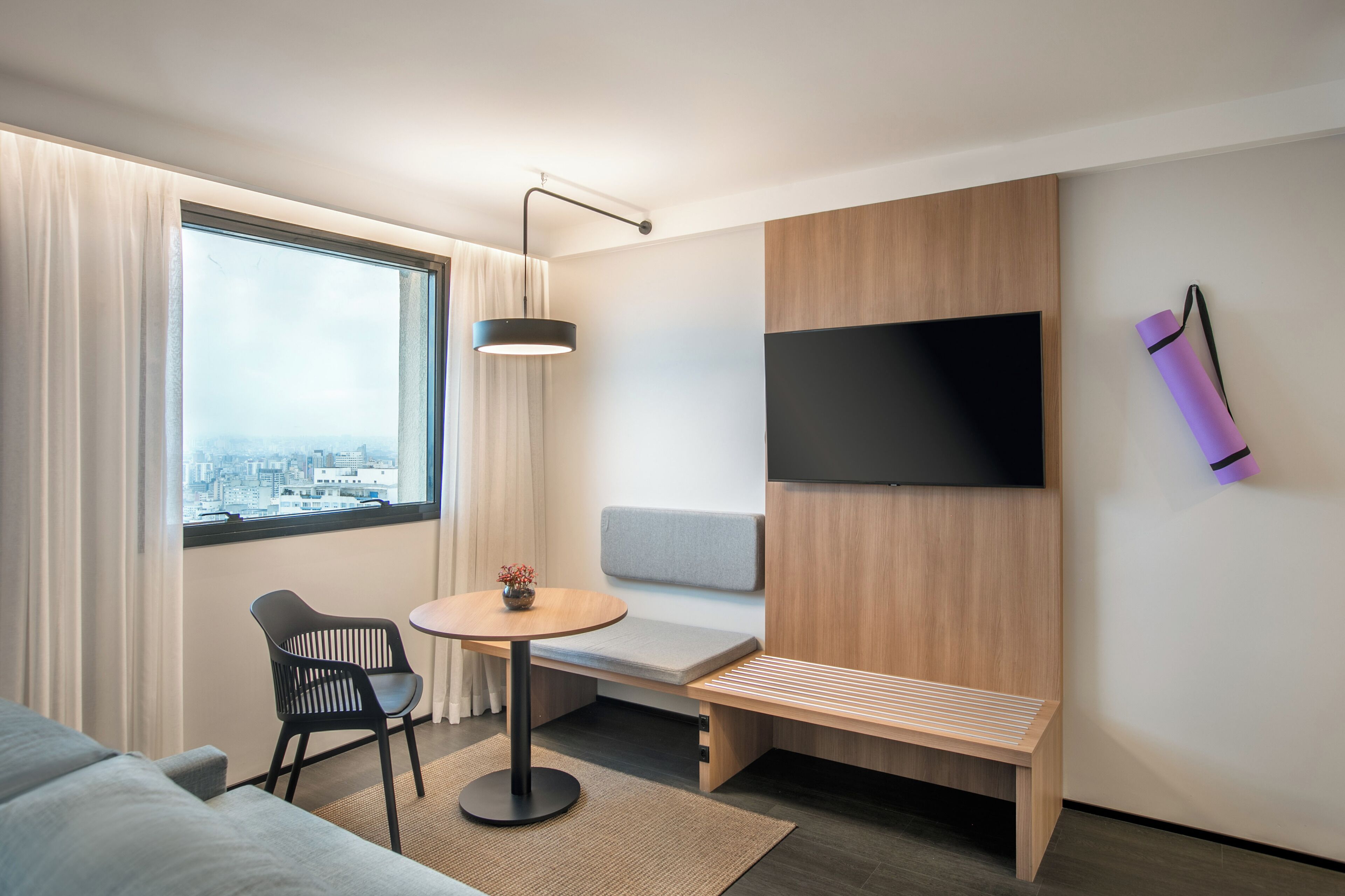 A modern hotel room with a large window, a sofa, a small round table, a bench, and a mounted TV.