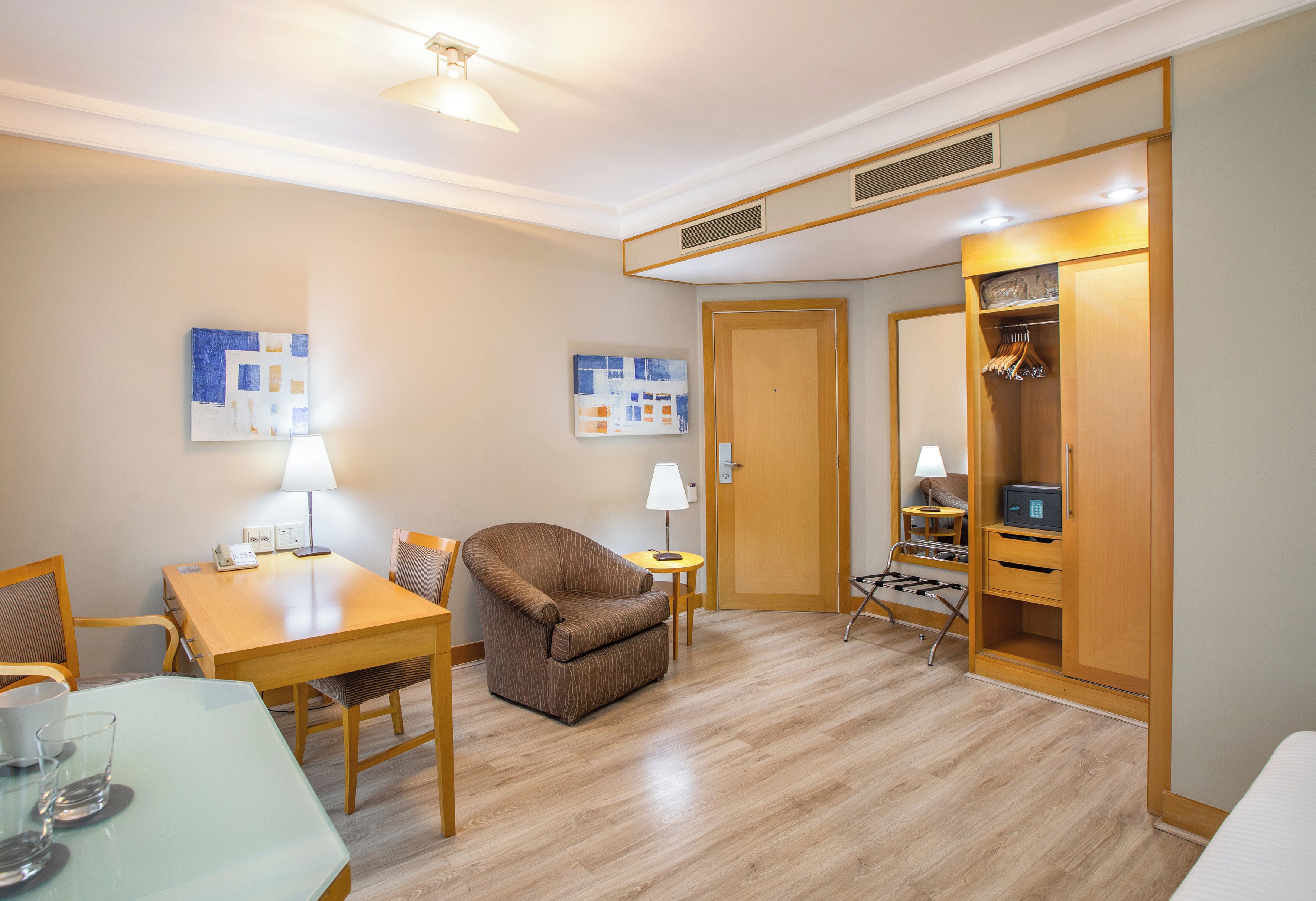 Ein Hotelzimmer mit Holzboden, einem Esstisch mit Stühlen, einer Couch, einem kleinen Beistelltisch mit Lampe, einem Kleiderschrank und zwei an der Wand hängenden Kunstwerken.