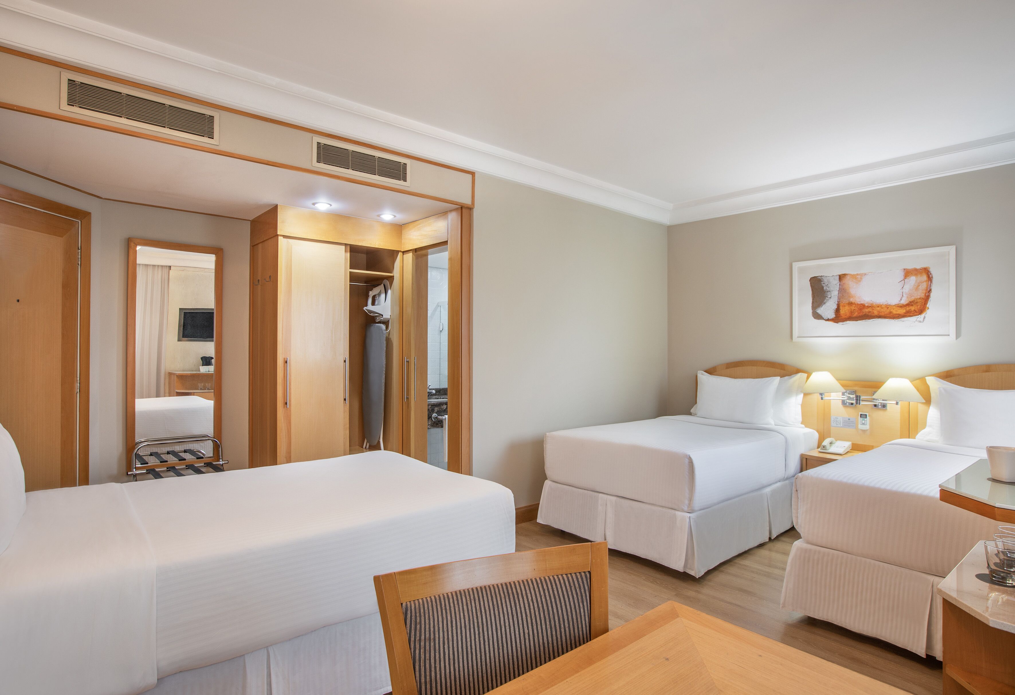 A hotel room with two beds, a wooden desk, a chair, a wardrobe, and a painting on the wall.