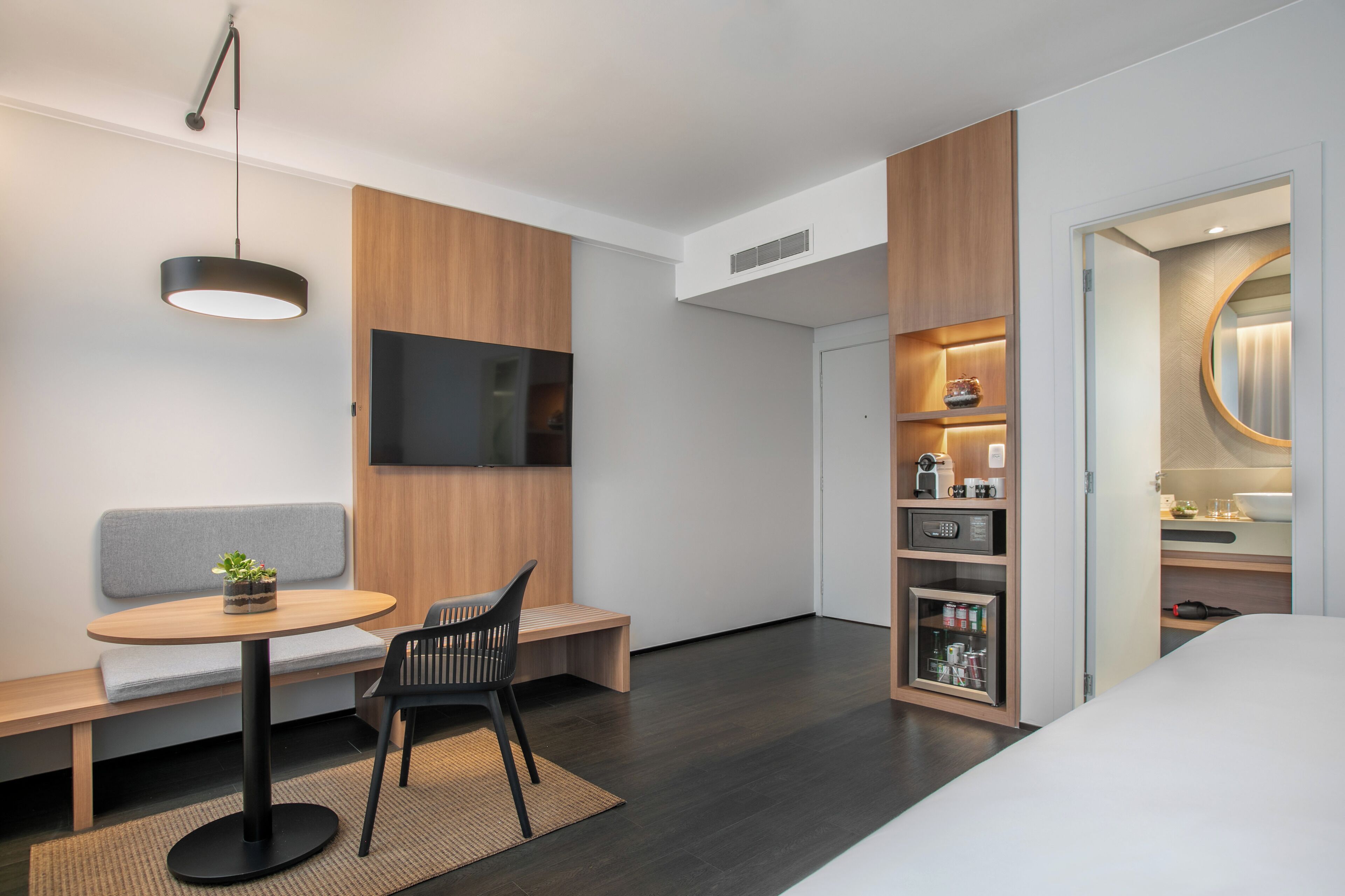 A modern hotel room with a wooden accent wall, a flat-screen TV, a small round table with a bench, a black chair, a bedside table with a lamp, and a built-in wardrobe with a mirror.