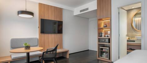 A modern hotel room with a wooden accent wall, a flat-screen TV, a small round table with a bench, a black chair, a bedside table with a lamp, and a built-in wardrobe with a mirror.