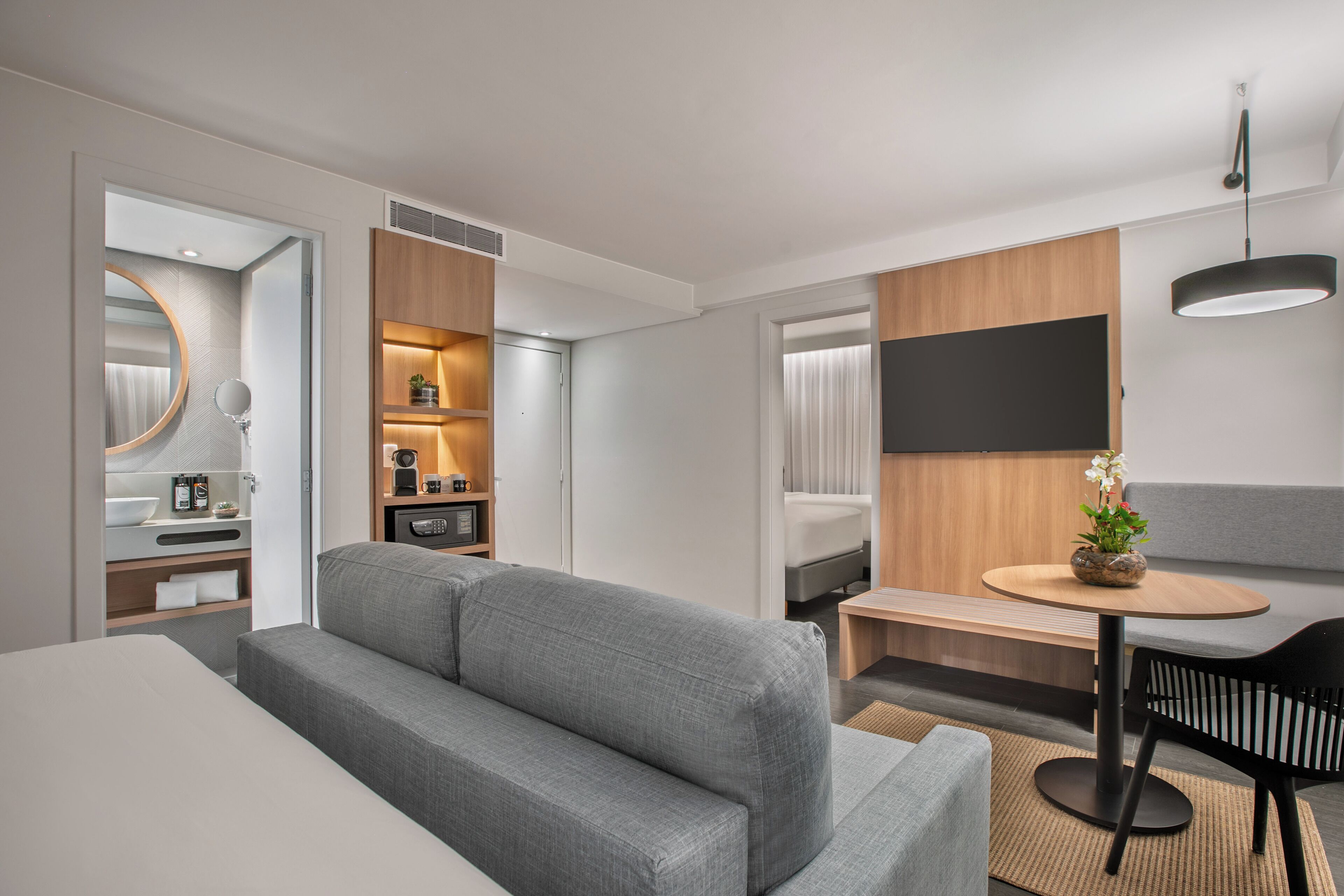 A modern hotel room with a grey sofa, a small round table with a potted plant, a black chair, a large mirror, and a flat-screen TV mounted on the wall.