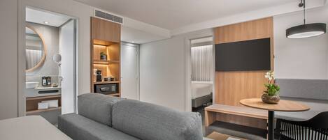 A modern hotel room with a grey sofa, a small round table with a potted plant, a black chair, a large mirror, and a flat-screen TV mounted on the wall.