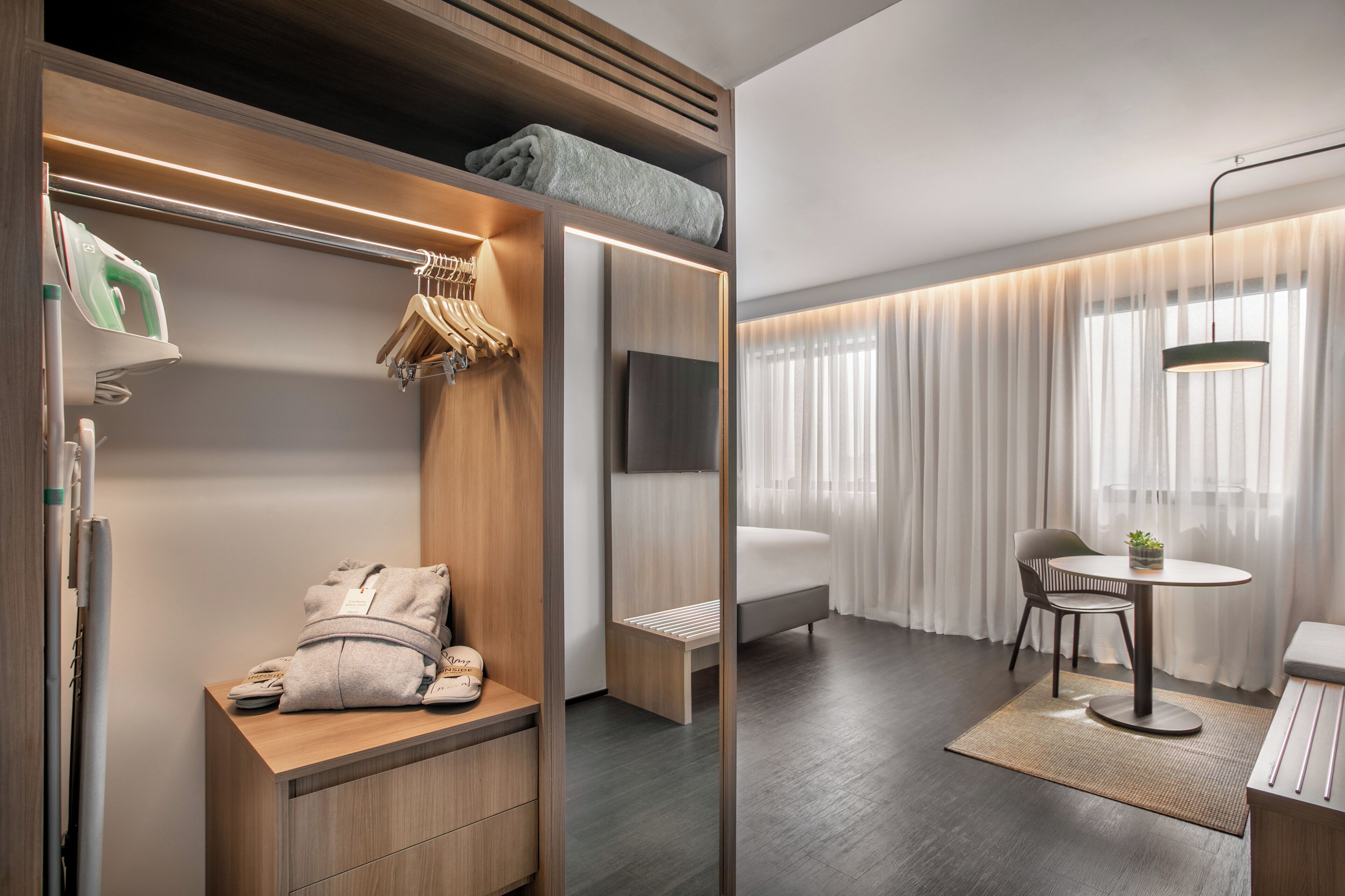 A modern hotel room with a wooden closet, a small seating area, a flat-screen TV, and a large window with sheer curtains.