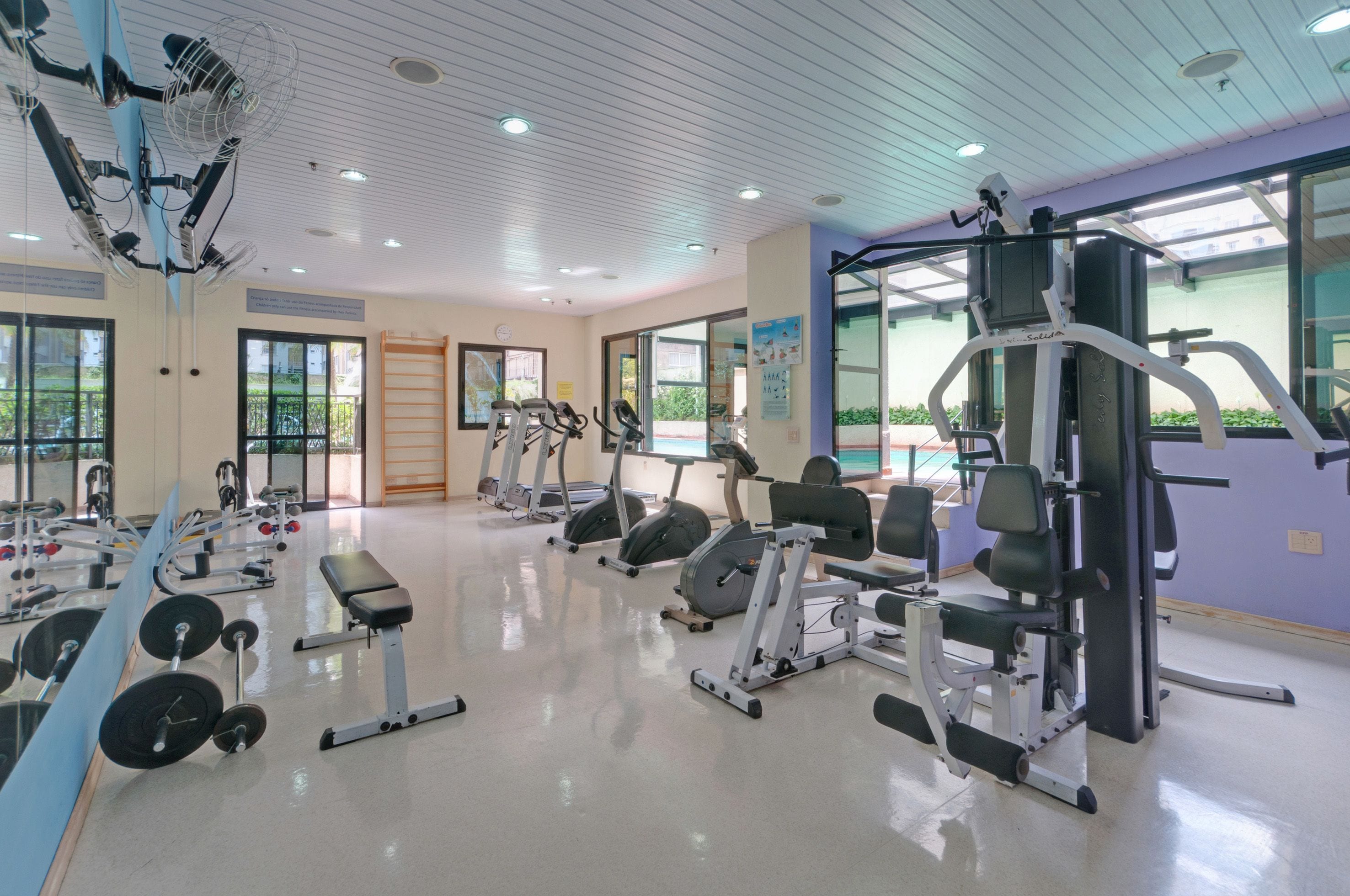 Un gimnasio con diverso equipo de ejercicio, amplias ventanas y un interior limpio y bien iluminado.