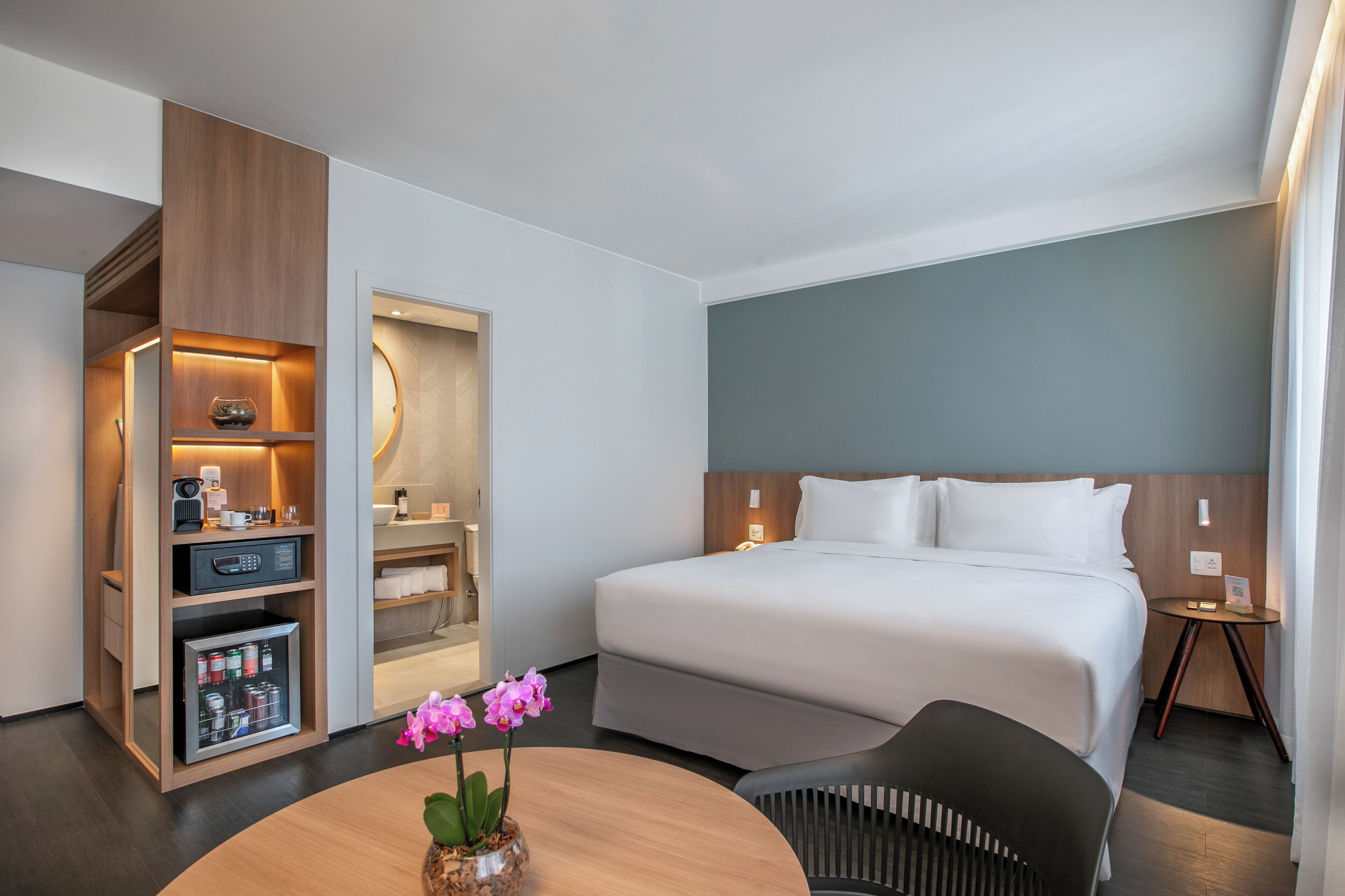 Una habitación de hotel moderna con una cama grande, un escritorio con computadora, un minibar y una pequeña mesa con flores.