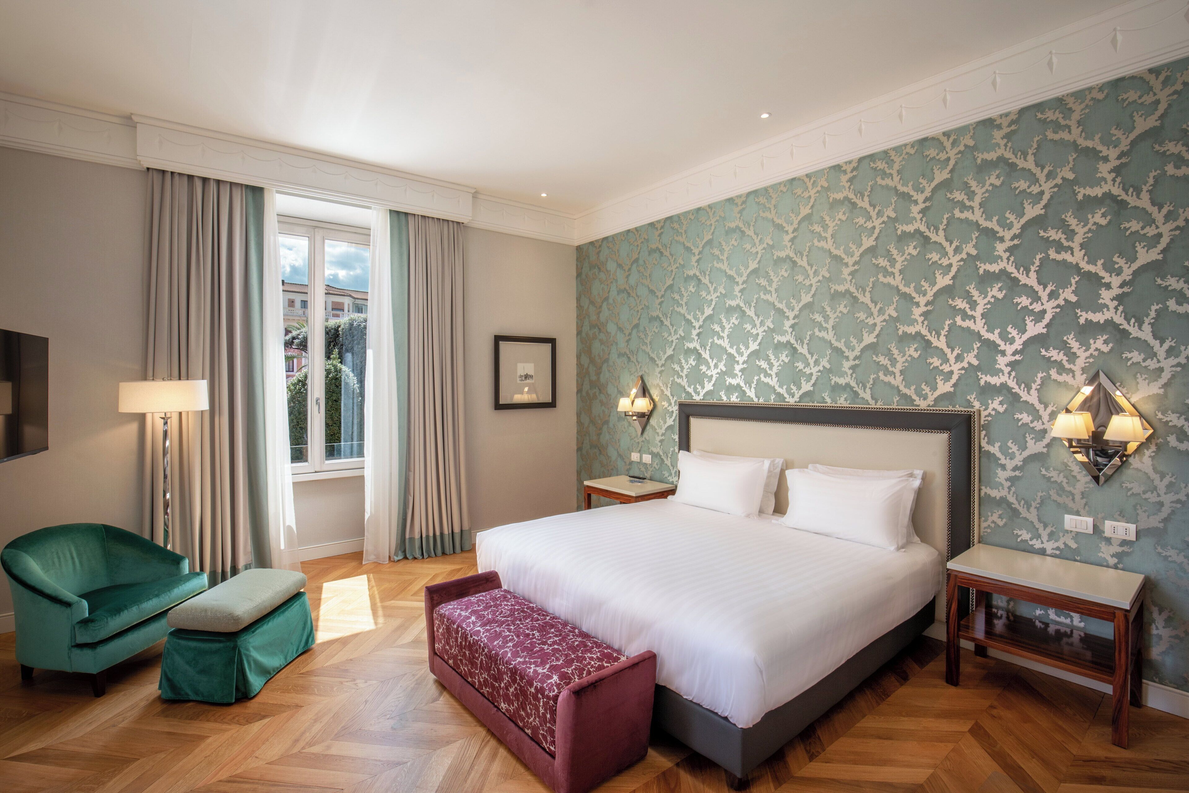 Quarto de hotel com uma cama grande, uma poltrona verde, um banco, uma mesinha de cabeceira e uma luminária na parede.