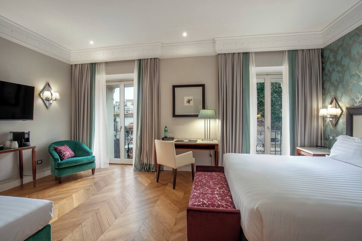 A hotel room with a large bed, a desk with a chair, a TV, and a balcony with curtains.