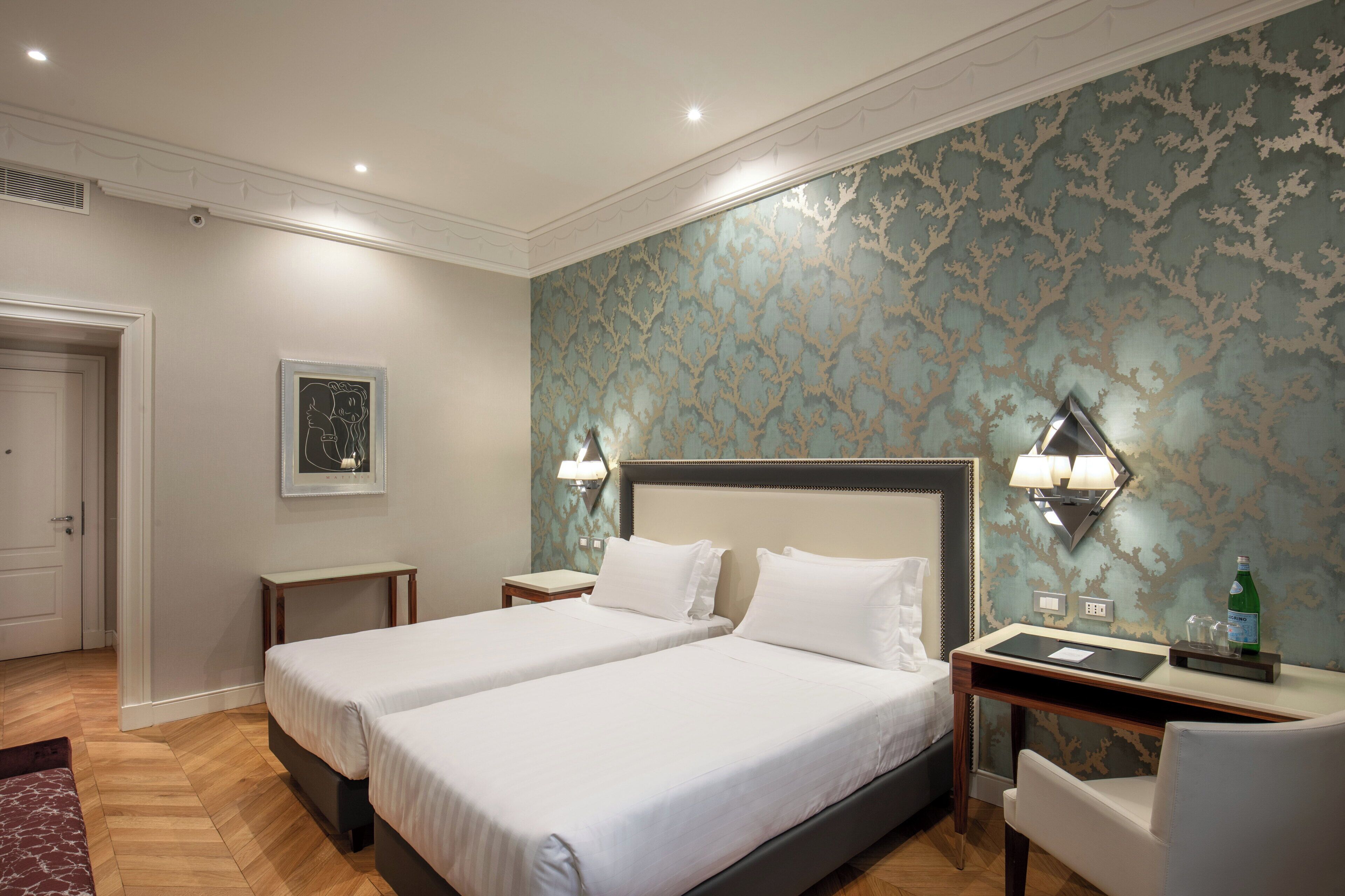 Une chambre d’hôtel avec deux lits, un bureau avec une lampe, une chaise et un mur recouvert d’un papier peint à motifs.