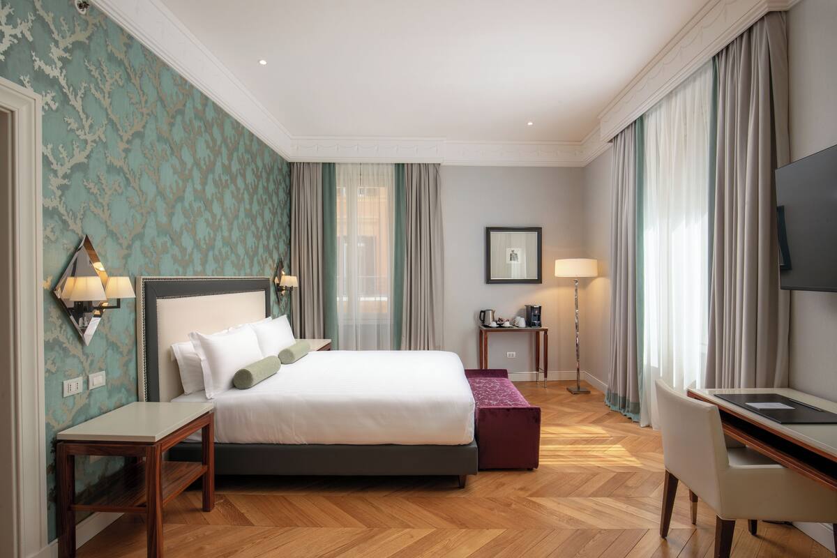 A hotel room with a bed, bedside table, lamp, desk, chair, and a television.