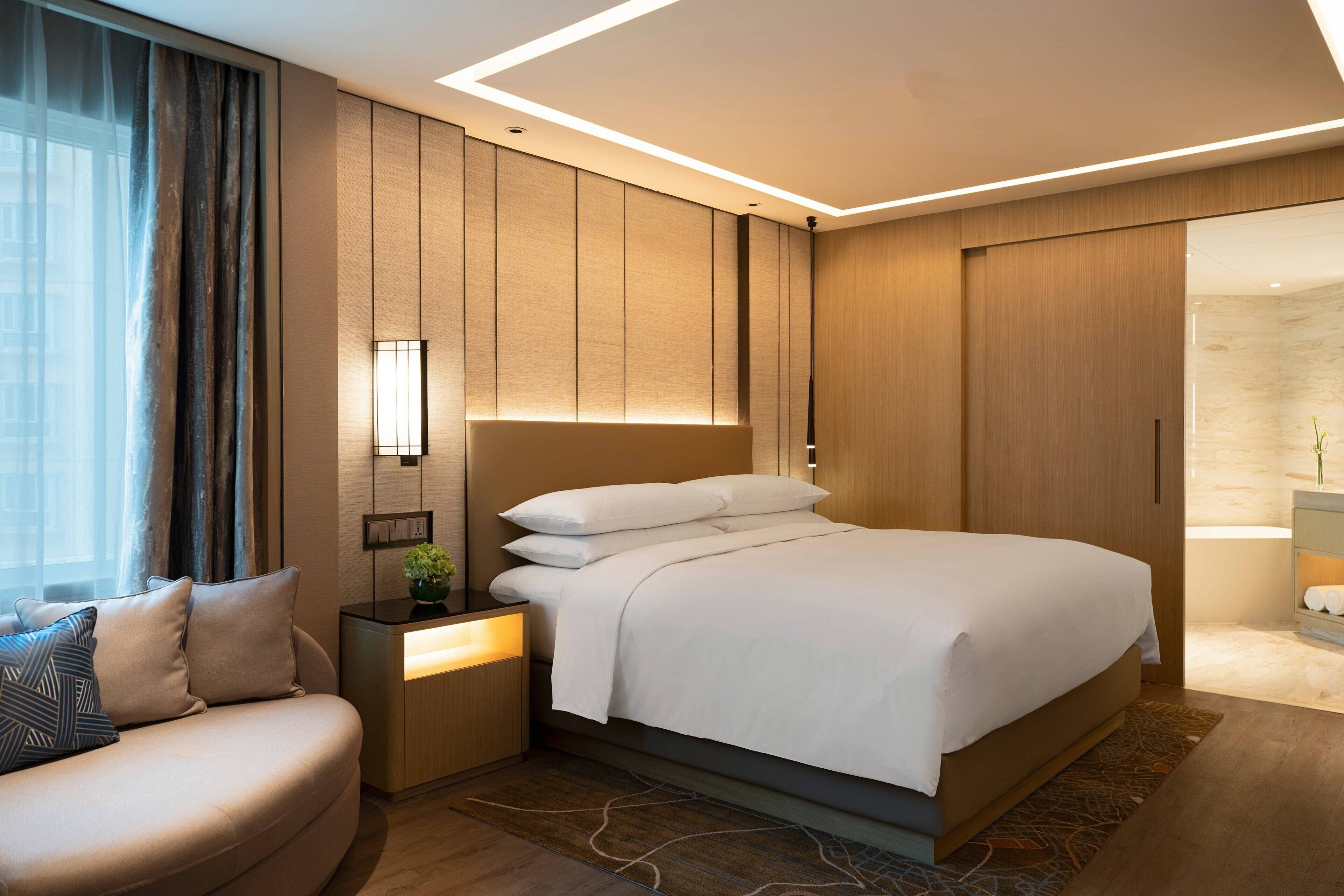 Habitación de hotel moderna con una cama grande, una mesita de noche con lámpara, un sofá y vistas al baño.