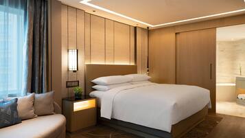 Habitación de hotel moderna con una cama grande, una mesita de noche con lámpara, un sofá y vistas al baño.