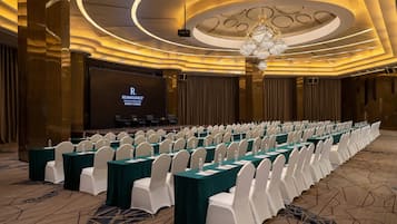 Une salle de conférence avec des rangées de tables et de chaises, un grand écran et un lustre.