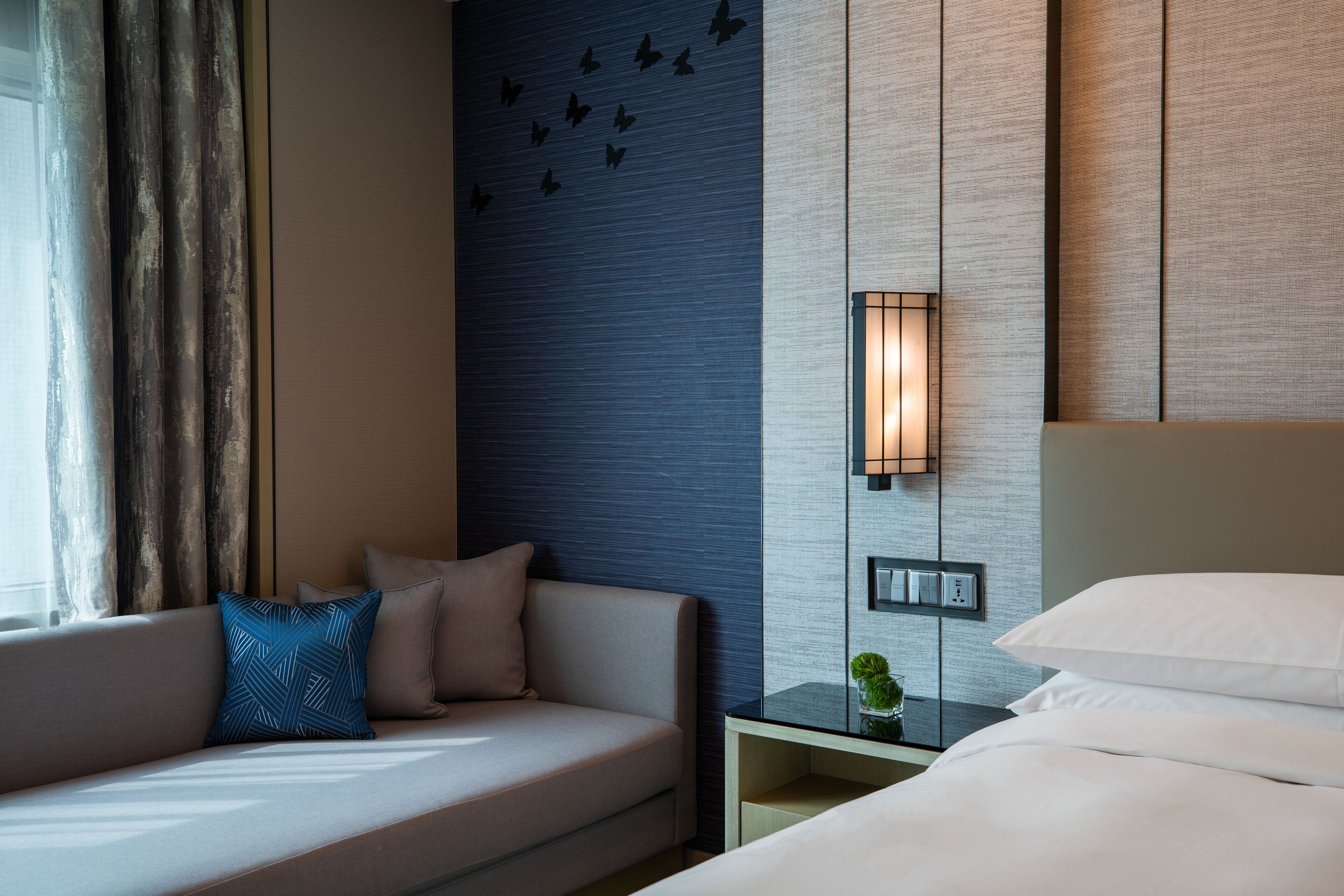 Una habitación de hotel moderna con un sofá, una cama, una mesita de noche con lámpara y una pared decorada con un adhesivo de mariposa.