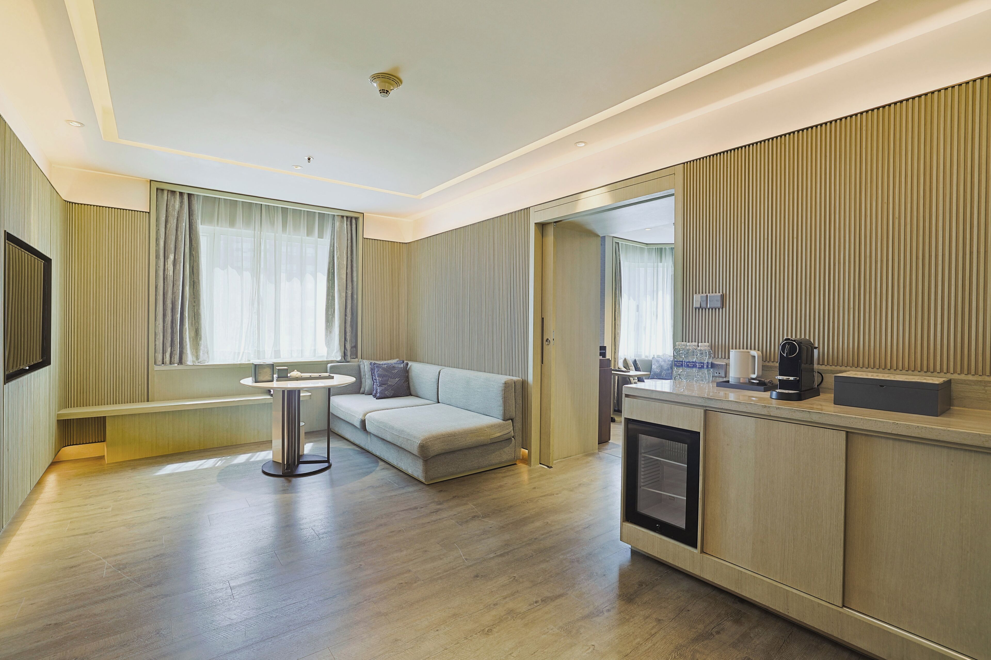 A modern hotel room with a sofa, a small round table, a coffee machine, and a view of the city.