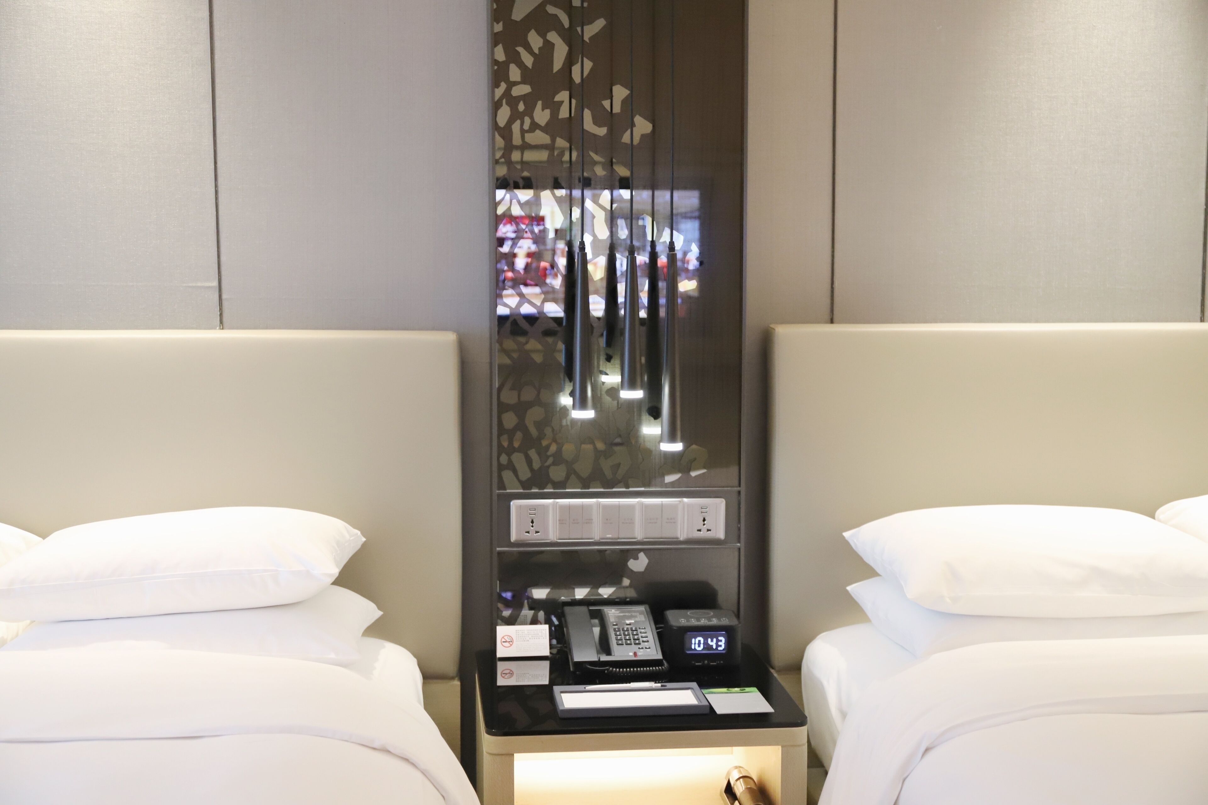 Une chambre d’hôtel avec deux lits, une table de chevet avec un téléphone et une horloge numérique, et un panneau décoratif avec des lumières.