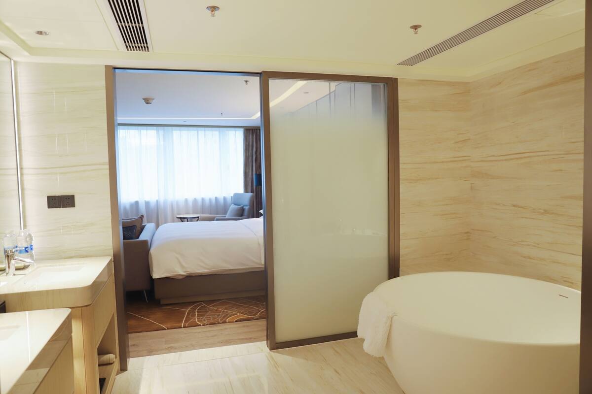 A modern hotel room with a bathroom, a bed, and a small sitting area.