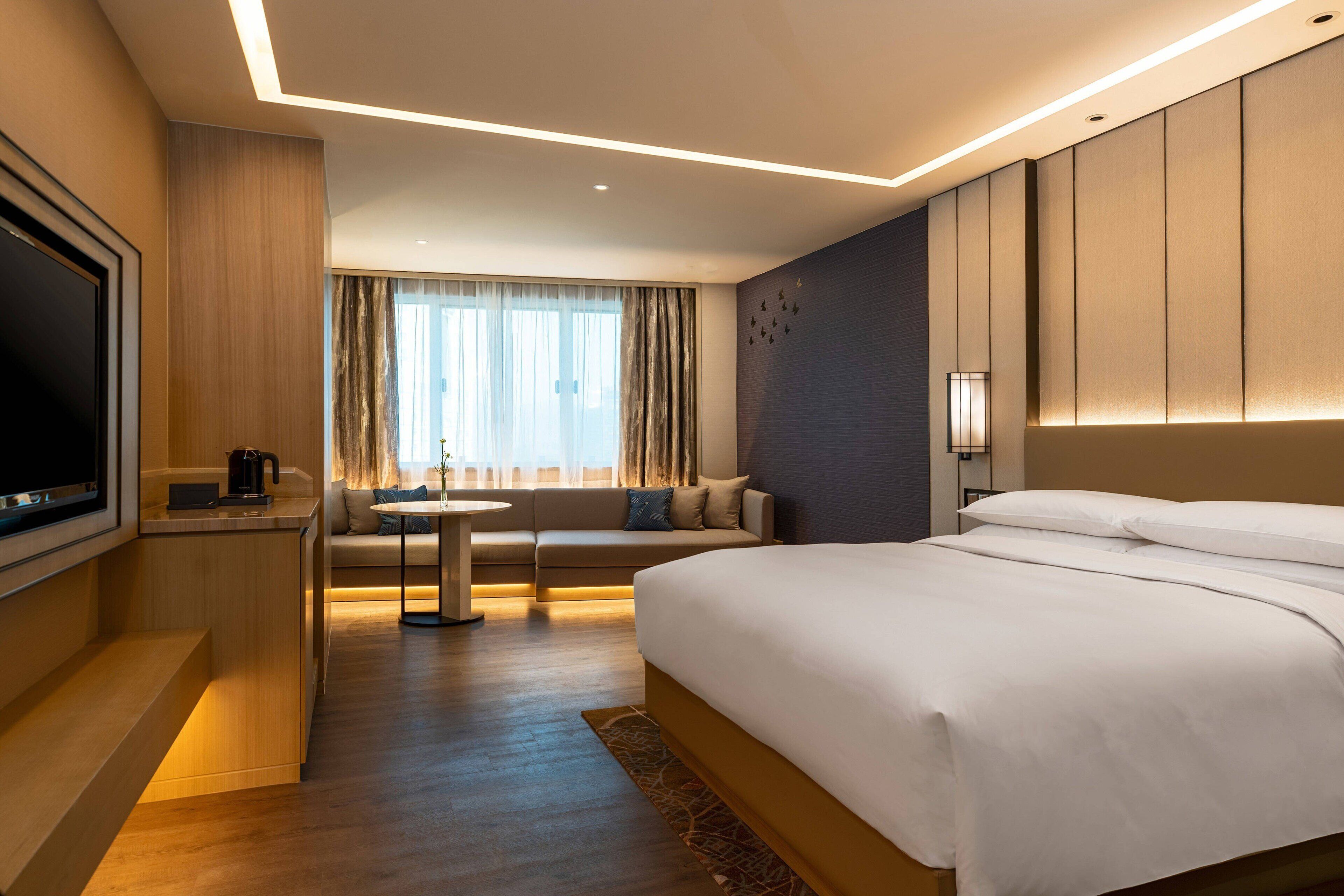 Habitación de hotel moderna con una cama grande, televisión, sofá, una mesita y una ventana con cortinas.