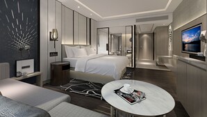 Una habitación de hotel moderna con una cama grande, una zona de estar con una mesa de centro, un televisor y vistas a la ciudad.