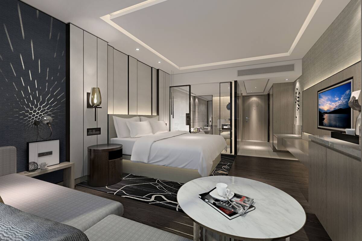 A modern hotel room with a large bed, a seating area with a coffee table, a TV, and a view of the cityscape.