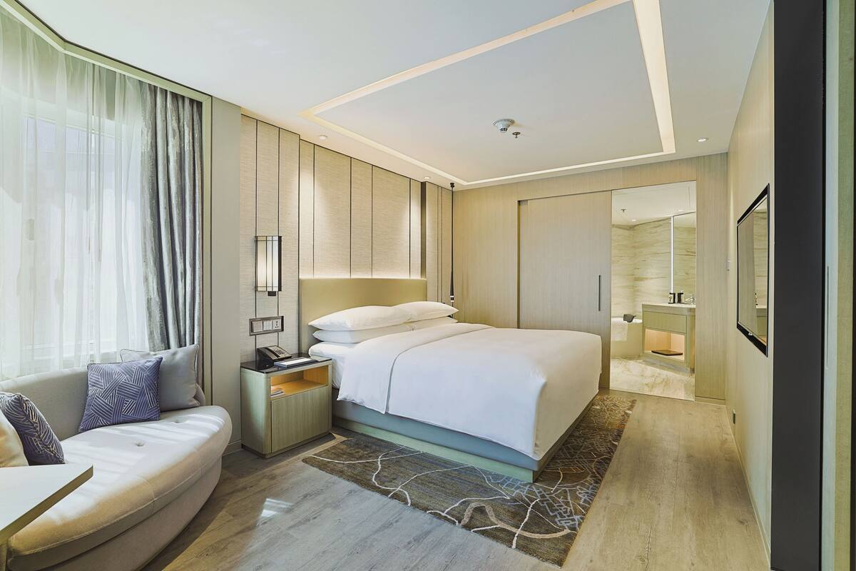 A modern hotel room with a bed, a sofa, a bedside table, a nightstand, a mirror, and a bathroom visible through an open door.