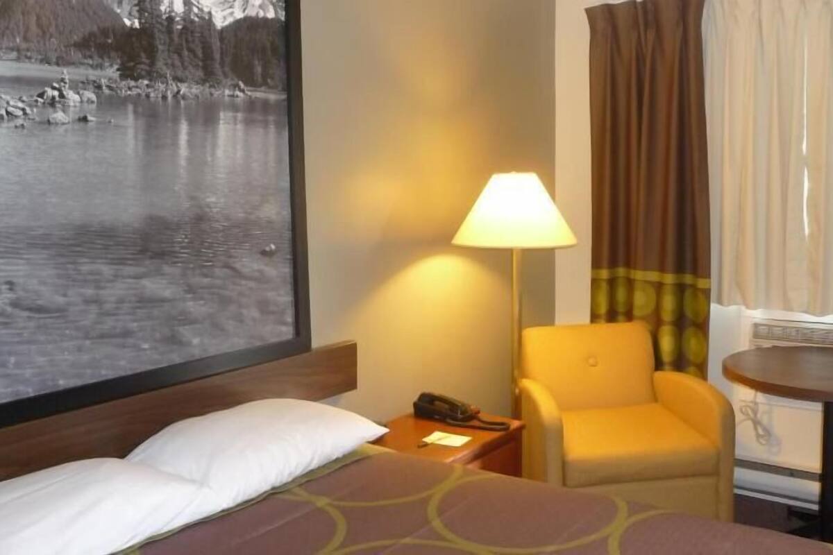 Une chambre d’hôtel avec un lit, une chaise, une lampe, une petite table et une grande fenêtre donnant sur un lac.