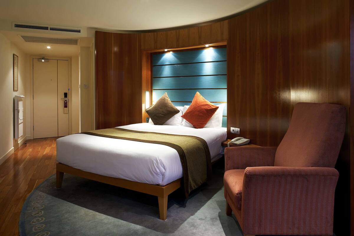 A hotel room with a bed, a chair, a bedside table with a phone, and a wall with a blue panel.