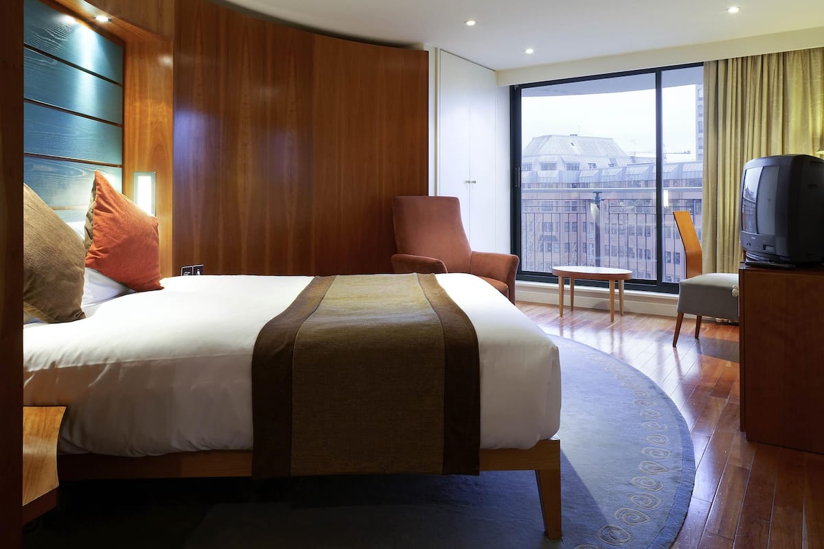 A hotel room with a large bed, a chair, a small table, a television, and a large window with a city view.