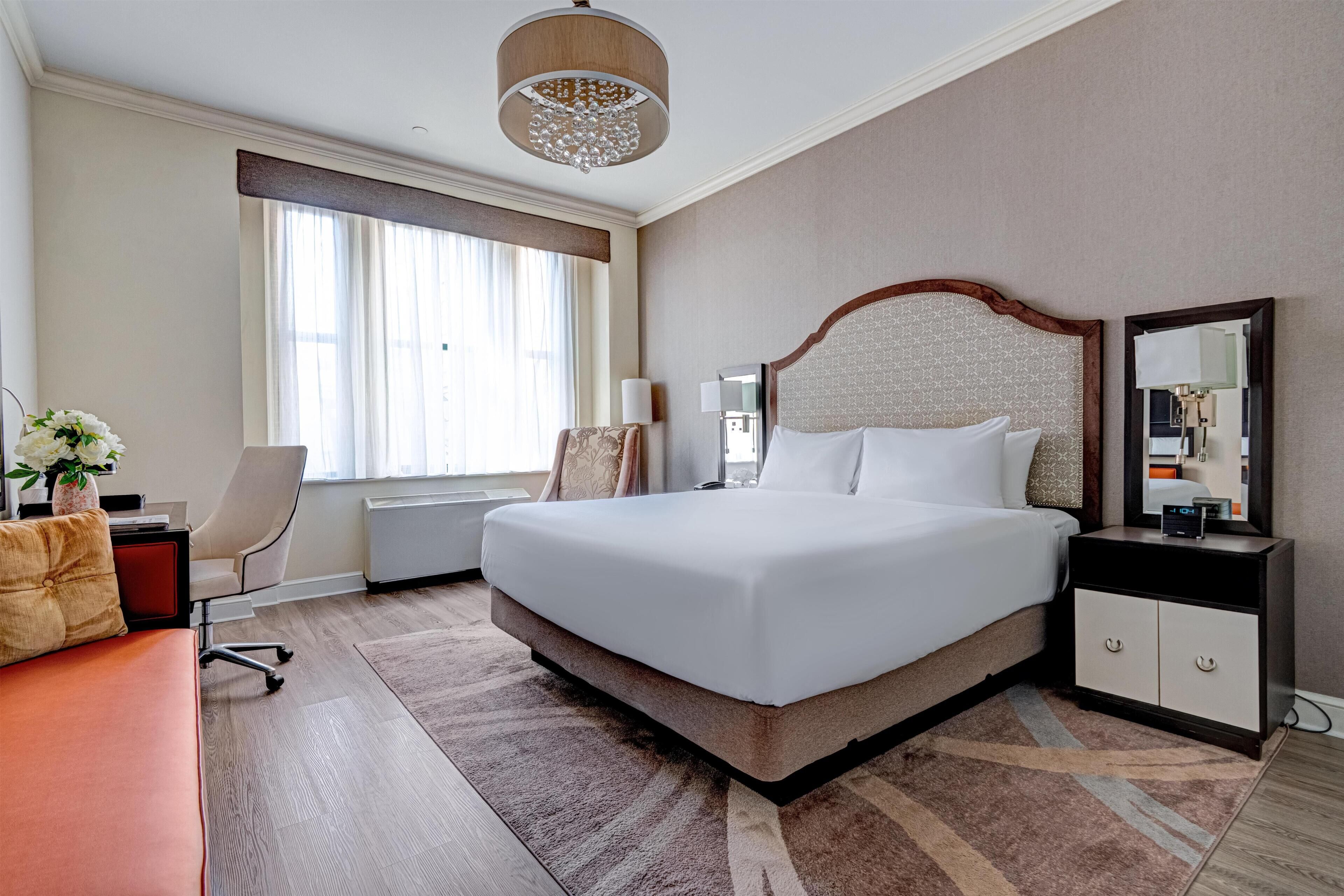 A hotel room with a large bed, a desk, a chair, a nightstand, a mirror, and a window with curtains.