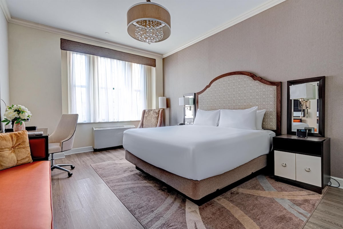 Quarto de hotel com uma cama grande, escrivaninha, cadeira, mesa de cabeceira, espelho e janela com cortinas.