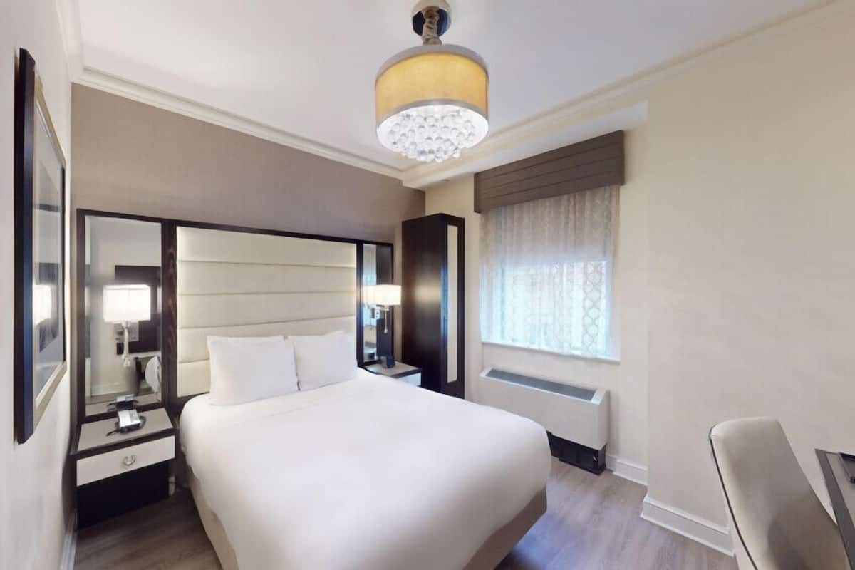 Quarto de hotel com uma cama grande, mesa de cabeceira, escrivaninha, cadeira, espelho, janela com cortinas e luminária no teto.