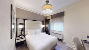 A hotel room with a large bed, a nightstand, a desk, a chair, a mirror, a window with curtains, and a ceiling light fixture.