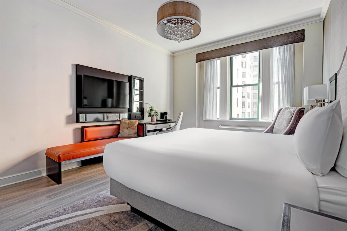Quarto de hotel moderno com cama grande, televisão de tela plana, um banco e uma janela ampla com vista.