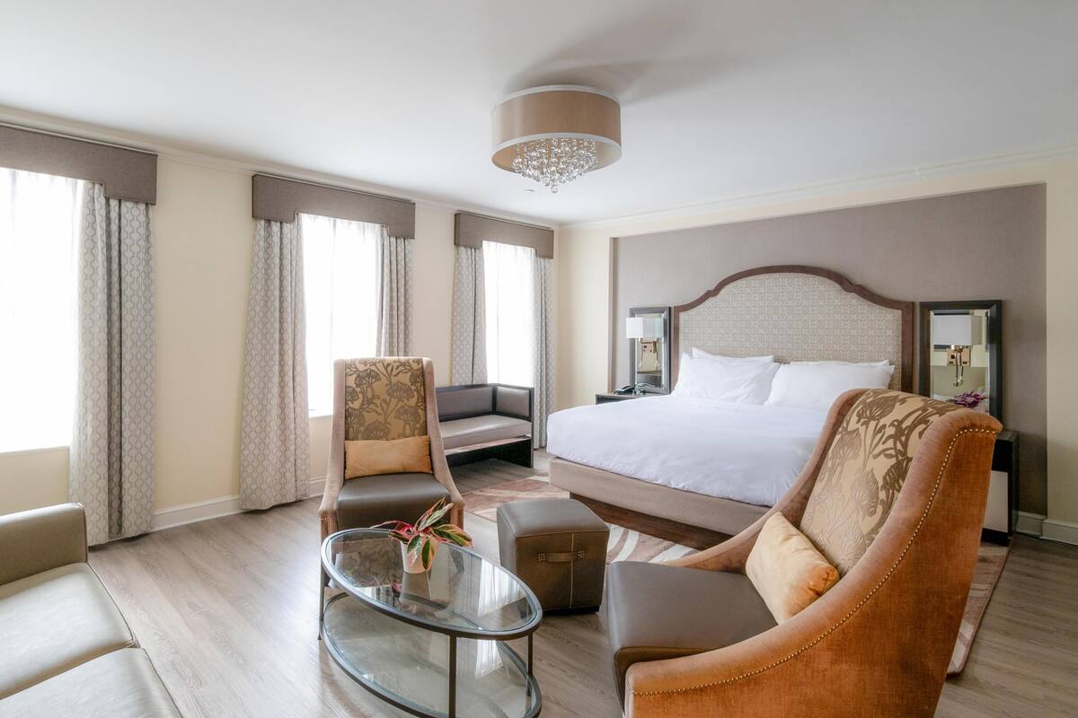 Quarto de hotel com uma cama grande, um sofá, uma cadeira, uma mesinha e um lustre.