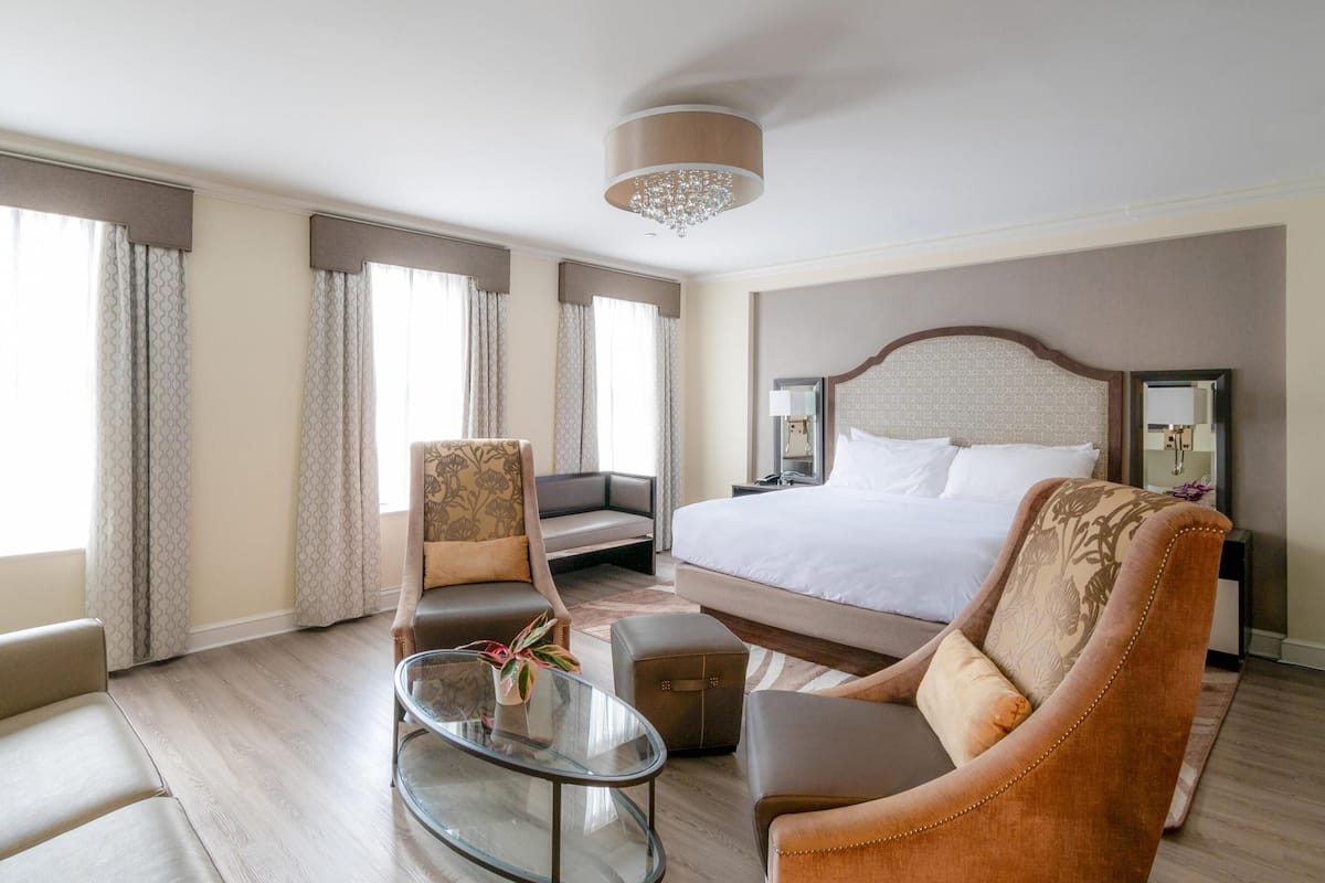 A hotel room with a large bed, a sofa, a chair, a small table, and a chandelier.