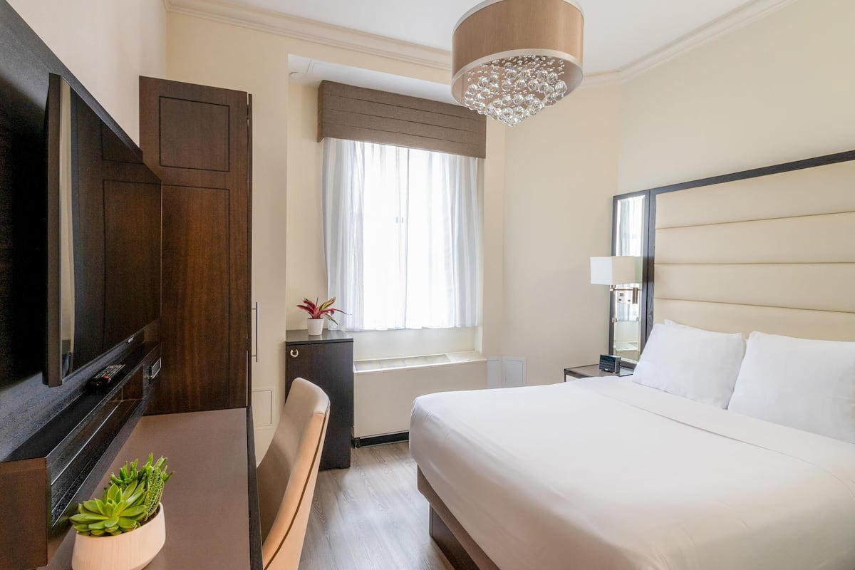 Quarto de hotel com cama, televisão, escrivaninha, janela com cortinas e um lustre.