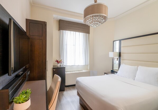 A hotel room with a bed, a television, a desk, a window with curtains, and a chandelier.
