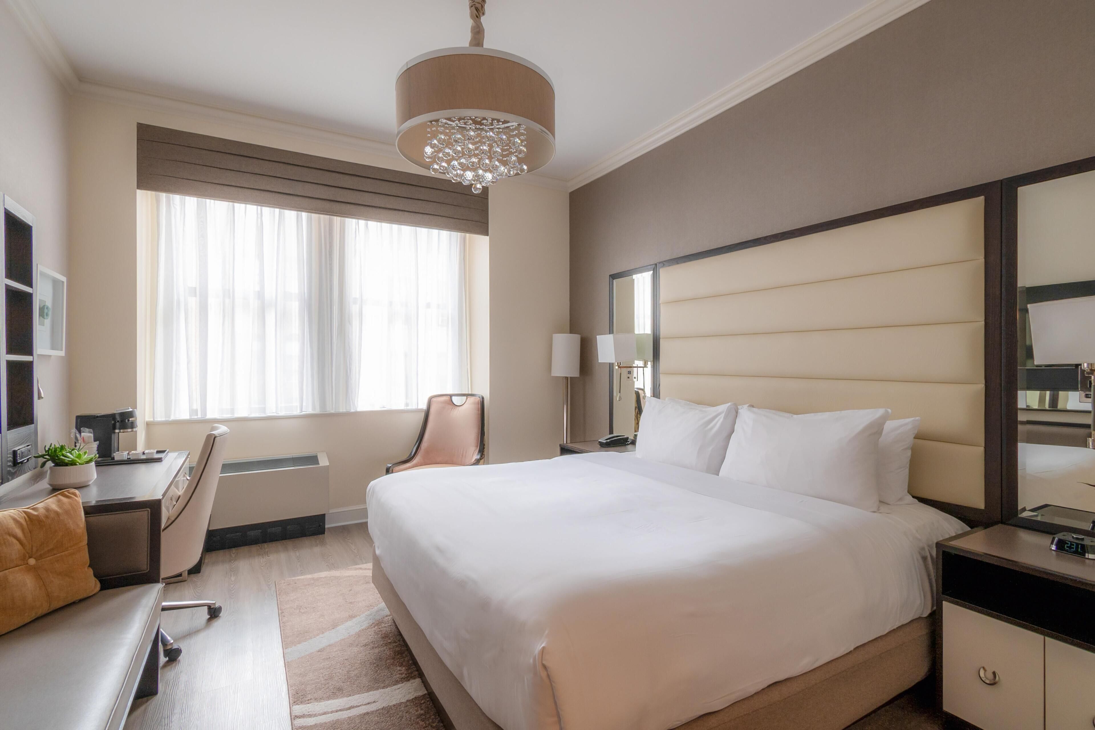 A hotel room with a large bed, a desk, a chair, a lamp, and a mirror.