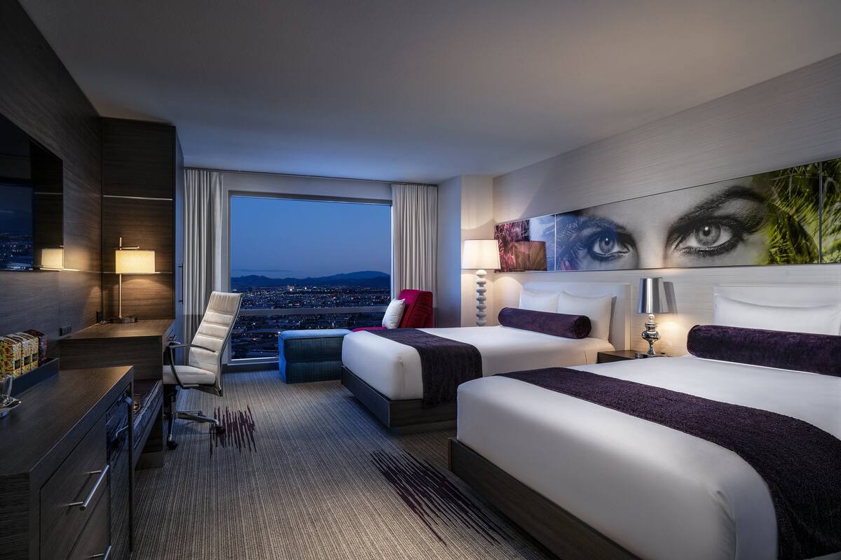 A hotel room with two beds, a desk, a chair, a large window with a city view, and a painting on the wall.