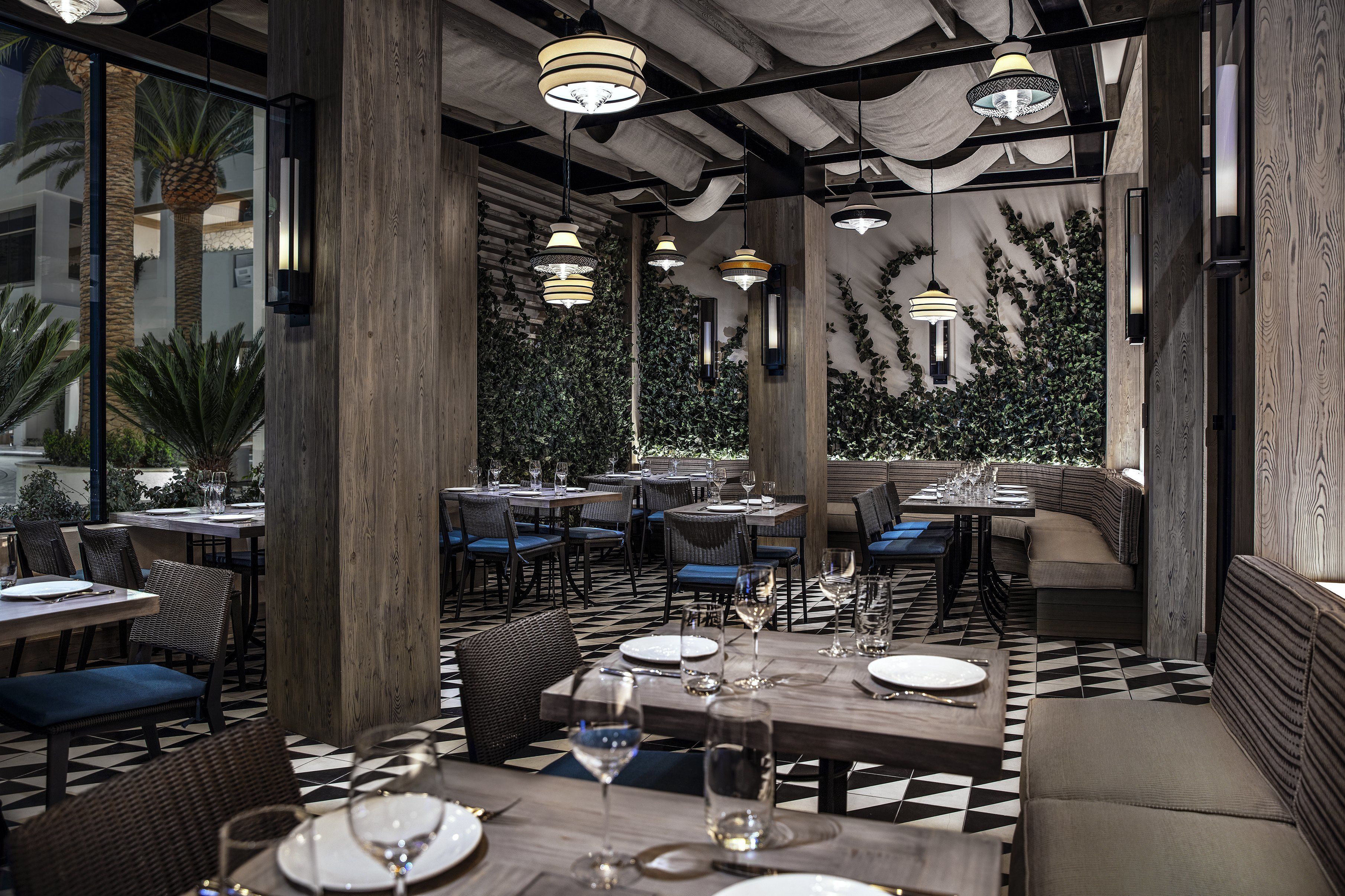 A restaurant with a checkered floor, wooden pillars, and hanging pendant lights. Tables are set with plates and glasses.