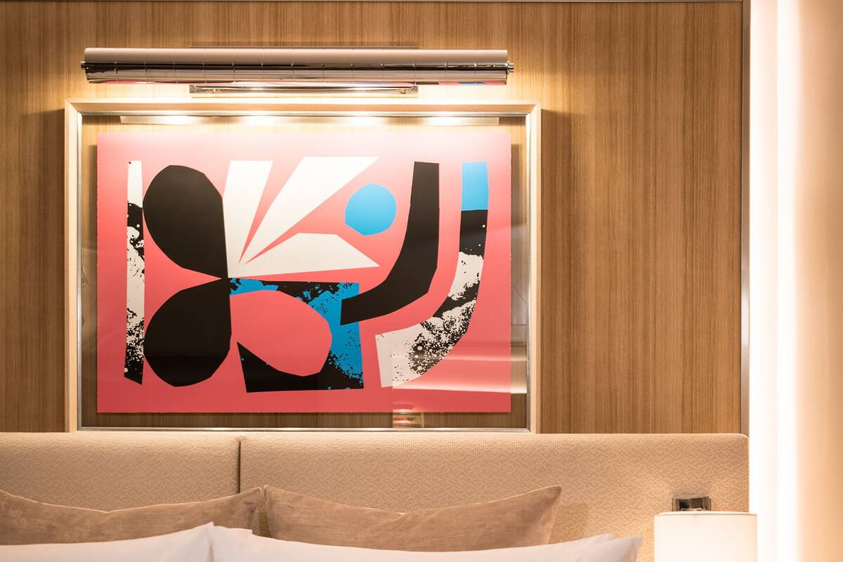 A hotel room with a large bed, a modern abstract painting on the wall, and a bedside lamp.