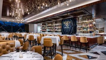 A spacious restaurant with round tables and leather booths, a bar area with a large selection of liquor bottles, and a modern chandelier.