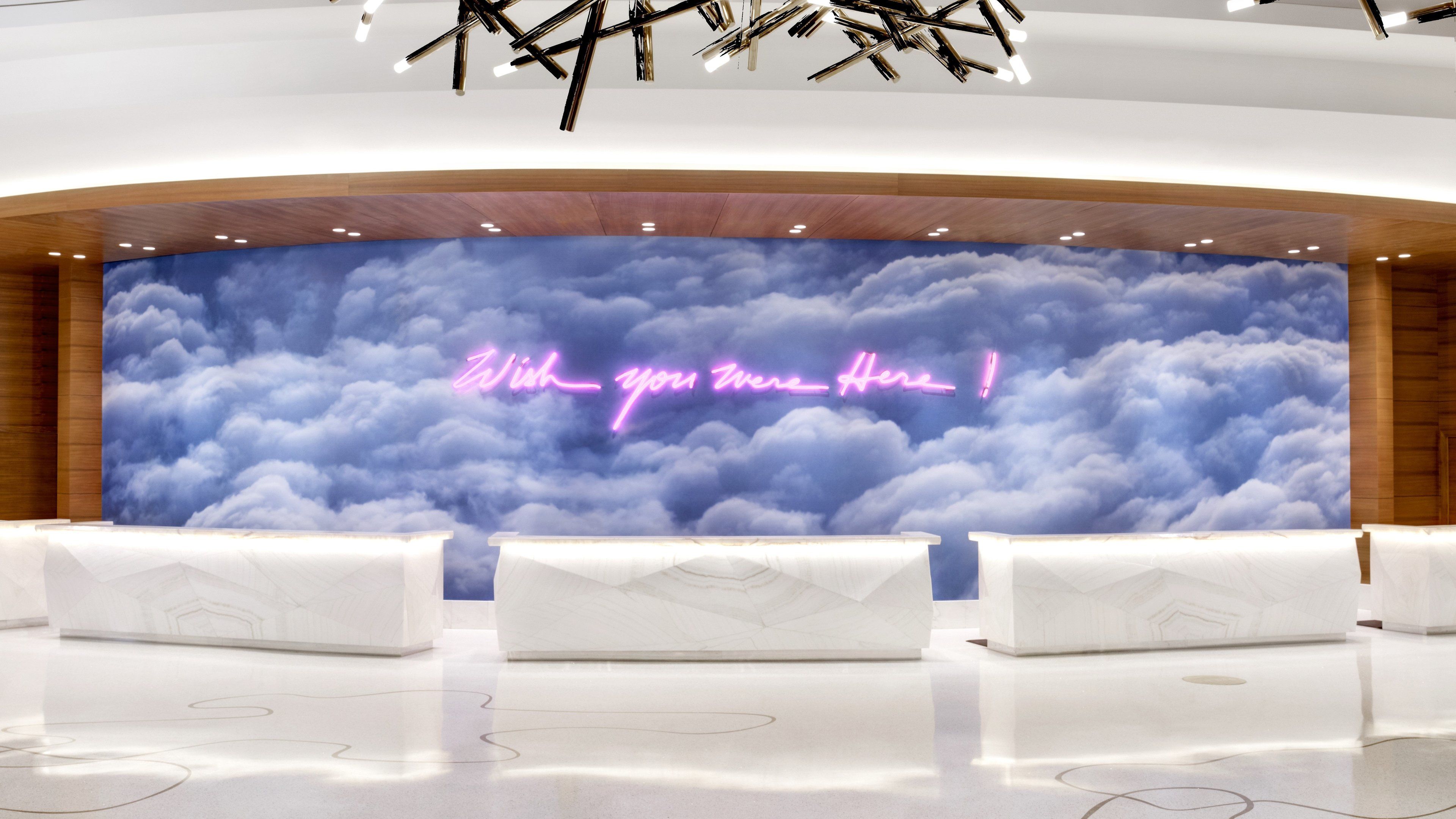 Uma área de recepção com um painel temático de nuvens e uma placa de neon com a frase 'Wish you were here'.