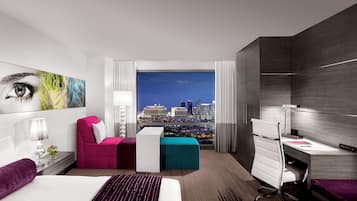 A modern hotel room with a city view, a large bed, a desk, and a comfortable seating area.